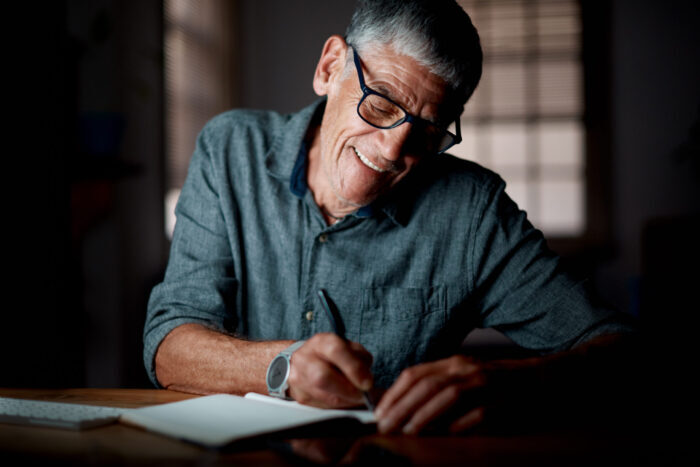Ein älterer Herr schreibt lächelnd in ein Notizbuch. Die autobiografische Rückschau mit einem Schreibprojekt abzuschließen, gibt die Möglichkeit, die Perlen der eigenen Biografie für sich selbst und andere Menschen zu bewahren.
